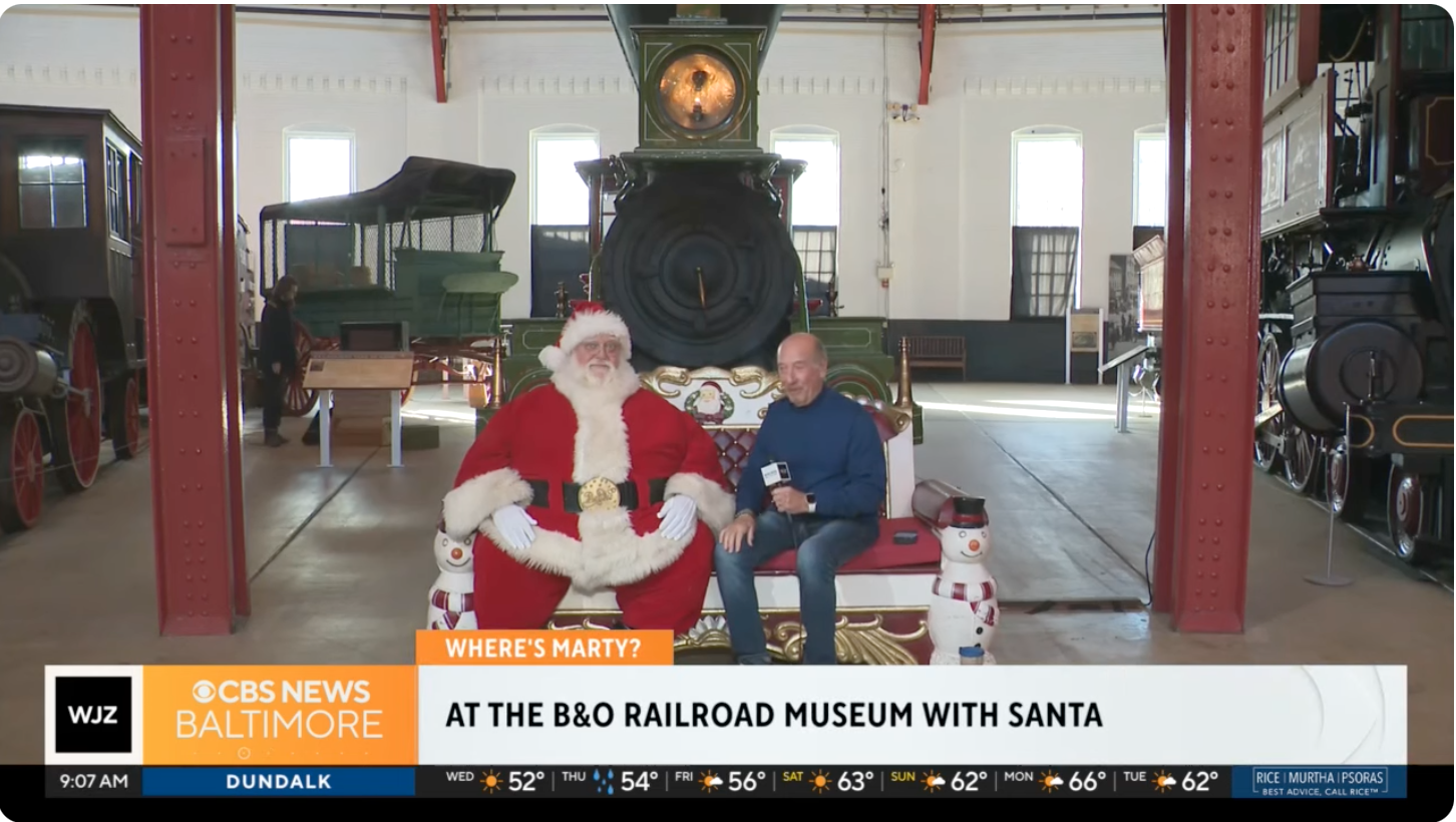 This is an image of Santa sitting on a bench. A reporter is sitting besides him. Behind them, you can see a red and green locomotive.